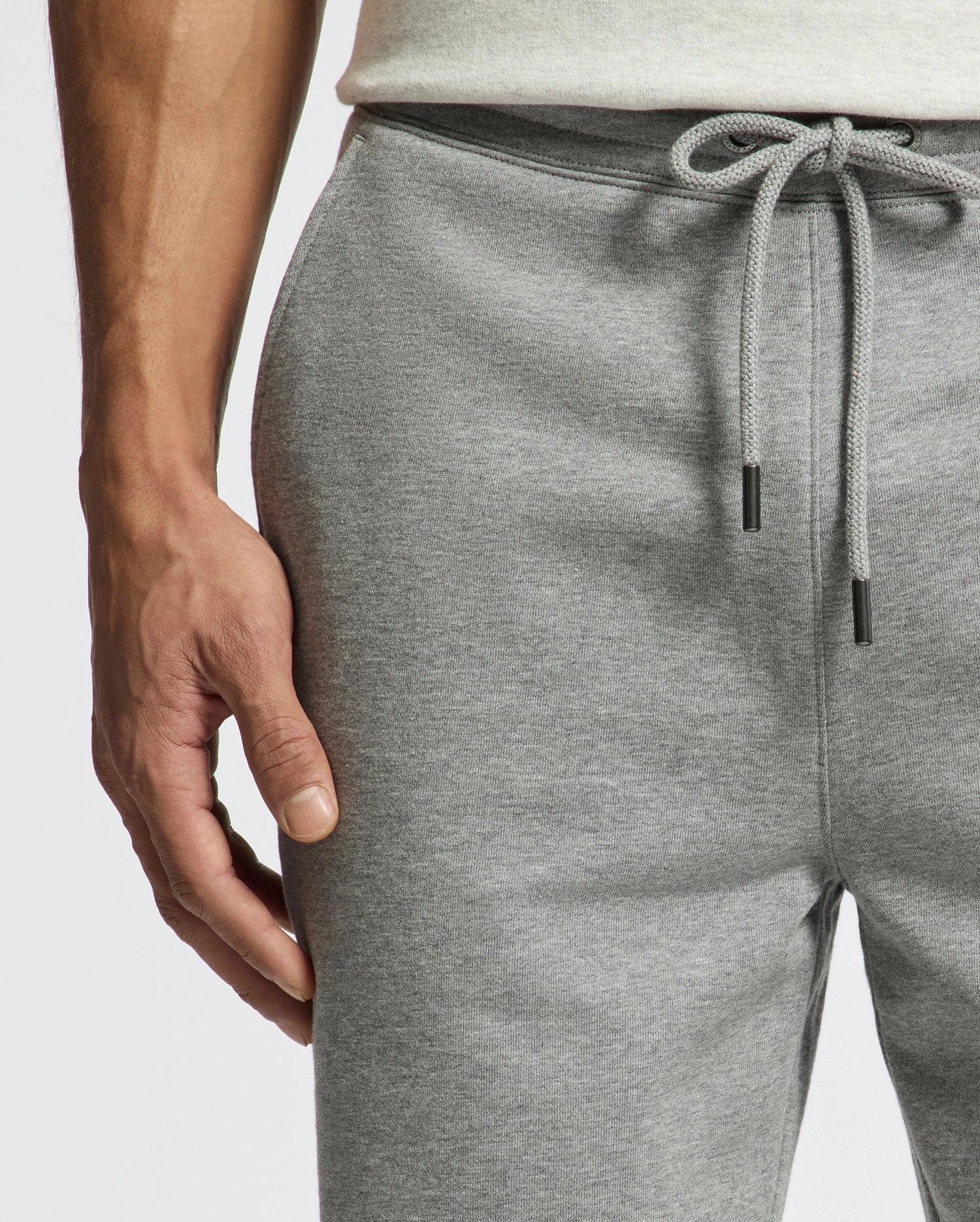 A person showcases the sleek Psycho Bunny MENS ELDORADO BRUSH BACK JOGGER - B6P285E200 in light gray with a drawstring, their left hand gracefully resting on the thigh, paired with a white or light-colored top against a plain white background.
