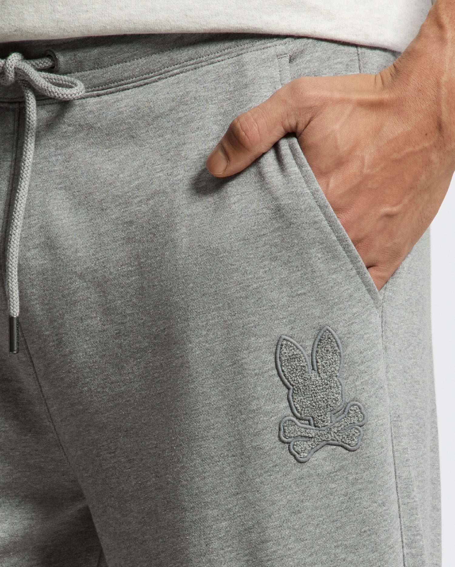 Close-up of someone wearing Psycho Bunny's Men's Eldorado Brush Back Jogger in gray, featuring an embroidered bunny with crossed bones. A drawstring waist provides all-day comfort, and a hand rests casually in the pocket.