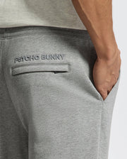 A close-up shows a hand in the pocket of sleek, stylish gray MENS ELDORADO BRUSH BACK JOGGER - B6P285E200 by Psycho Bunny, with "PSYCHO BUNNY" embroidered above the pocket.