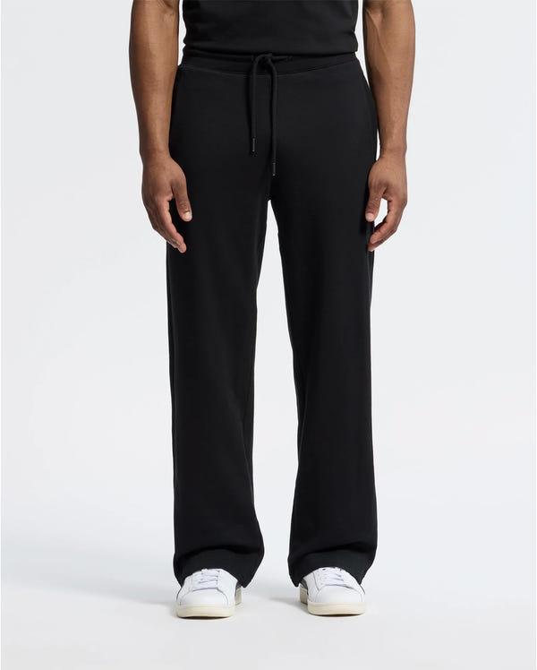 A person stands against a plain white background wearing black Psycho Bunny MENS LYNX WIDE LEG CLASSIC FRENCH TERRY SWEATPANT (B6P379E200) and white sneakers, showcasing the relaxed fit of the French terry fabric. Only the lower half is visible.