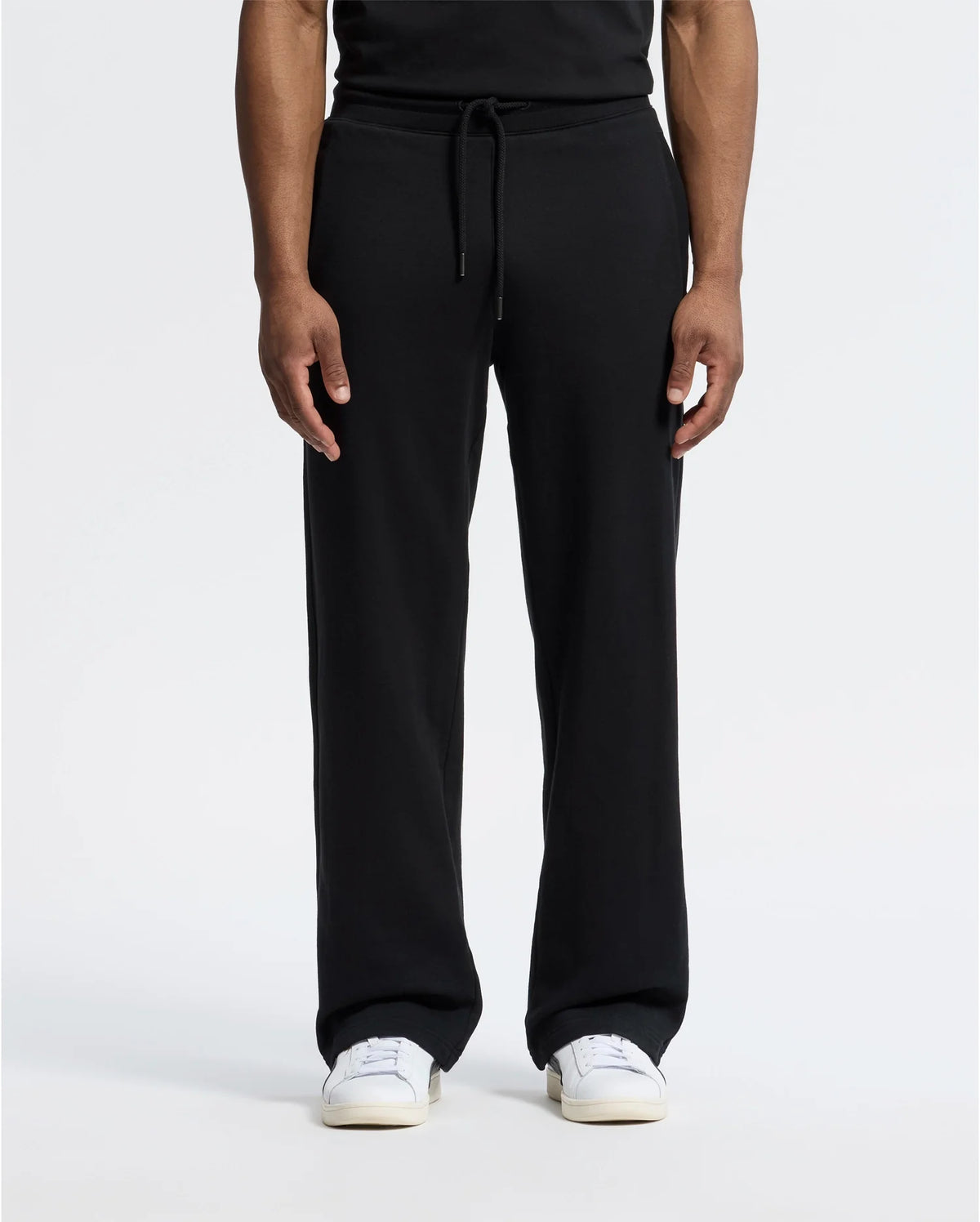 A person stands against a plain white background wearing black Psycho Bunny MENS LYNX WIDE LEG CLASSIC FRENCH TERRY SWEATPANT (B6P379E200) and white sneakers, showcasing the relaxed fit of the French terry fabric. Only the lower half is visible.