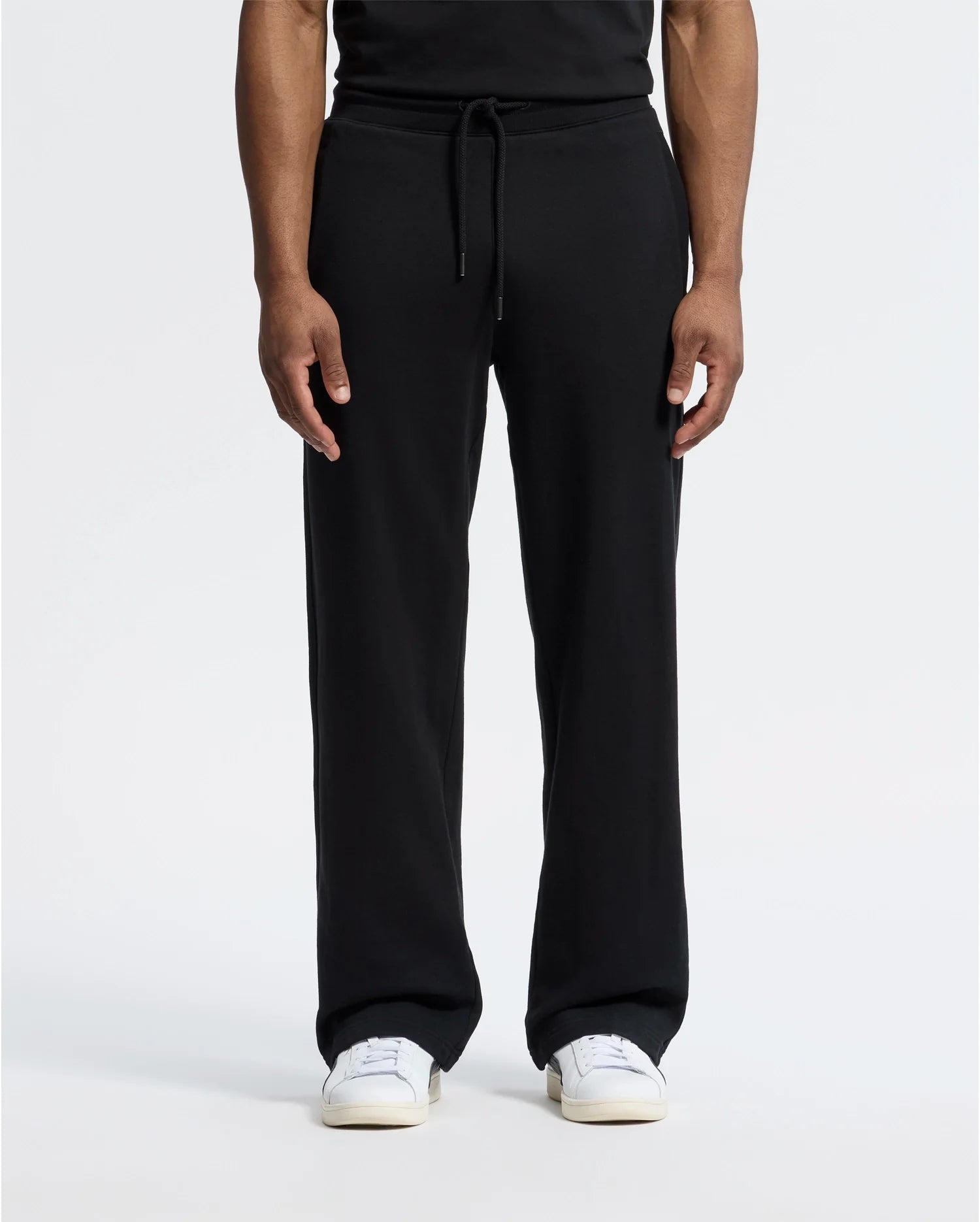 A person stands against a plain white background wearing black Psycho Bunny MENS LYNX WIDE LEG CLASSIC FRENCH TERRY SWEATPANT (B6P379E200) and white sneakers, showcasing the relaxed fit of the French terry fabric. Only the lower half is visible.