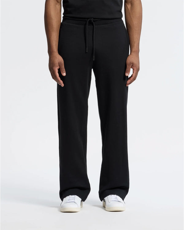 A person stands against a plain white background wearing black Psycho Bunny MENS LYNX WIDE LEG CLASSIC FRENCH TERRY SWEATPANT (B6P379E200) and white sneakers, showcasing the relaxed fit of the French terry fabric. Only the lower half is visible.