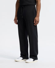 A person in Psycho Bunny's black MENS LYNX WIDE LEG CLASSIC FRENCH TERRY SWEATPANT - B6P379E200 with a relaxed fit and drawstring waist, paired with a black shirt and white sneakers, stands against a plain white background.