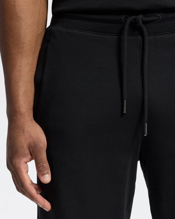 A person wearing Psycho Bunny's MENS LYNX WIDE LEG CLASSIC FRENCH TERRY SWEATPANT - B6P379E200 stands against a light background. Only the lower torso and hand are visible, with the hand resting near the pocket, subtly displaying an embroidered logo.
