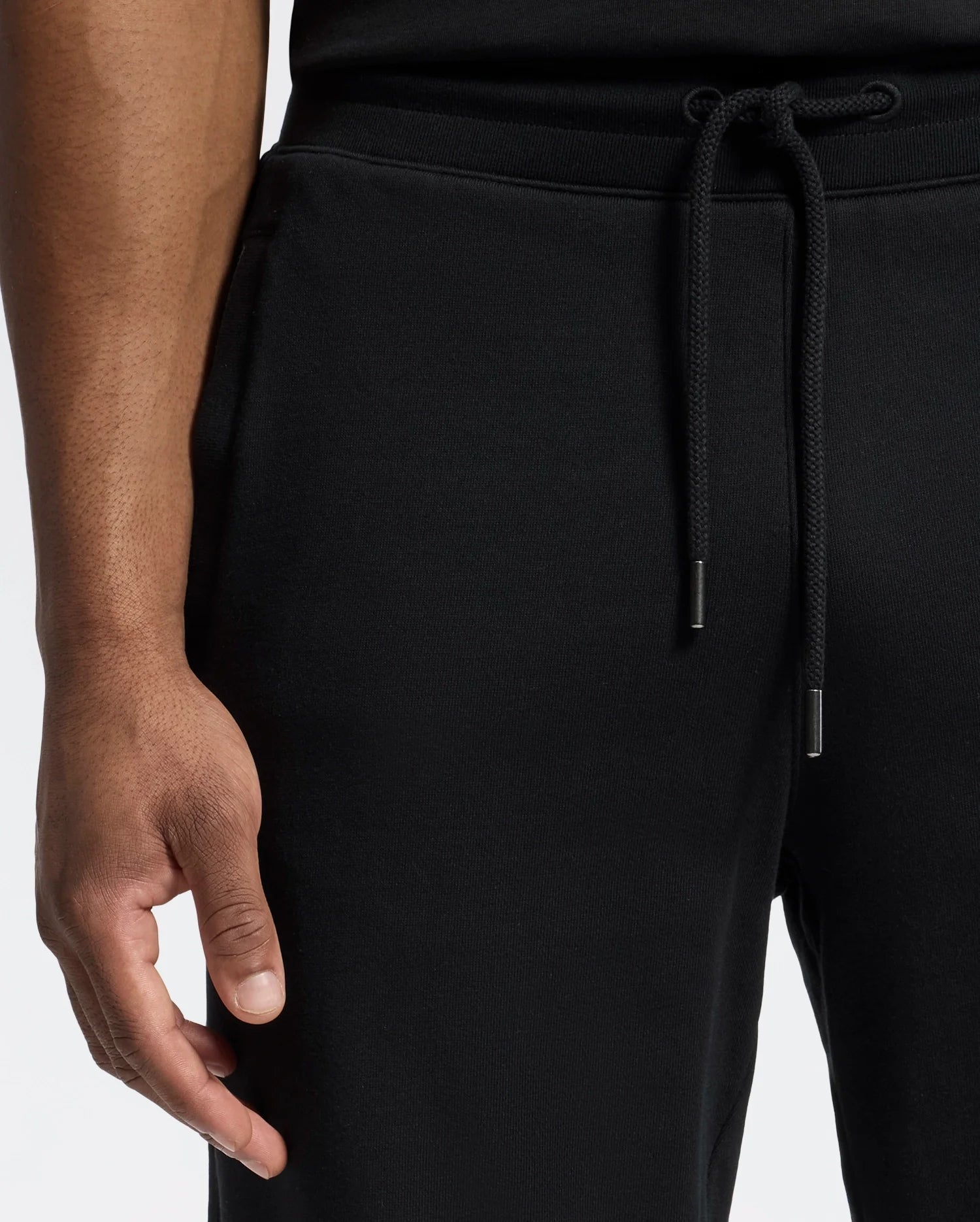 A person wearing Psycho Bunny's MENS LYNX WIDE LEG CLASSIC FRENCH TERRY SWEATPANT - B6P379E200 stands against a light background. Only the lower torso and hand are visible, with the hand resting near the pocket, subtly displaying an embroidered logo.