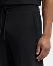 A close-up features a person wearing Psycho Bunny's MENS LYNX WIDE LEG CLASSIC FRENCH TERRY SWEATPANT (B6P379E200) in black with a drawstring and side pockets, an embroidered logo near the pocket, and their right hand relaxed against a plain white backdrop.