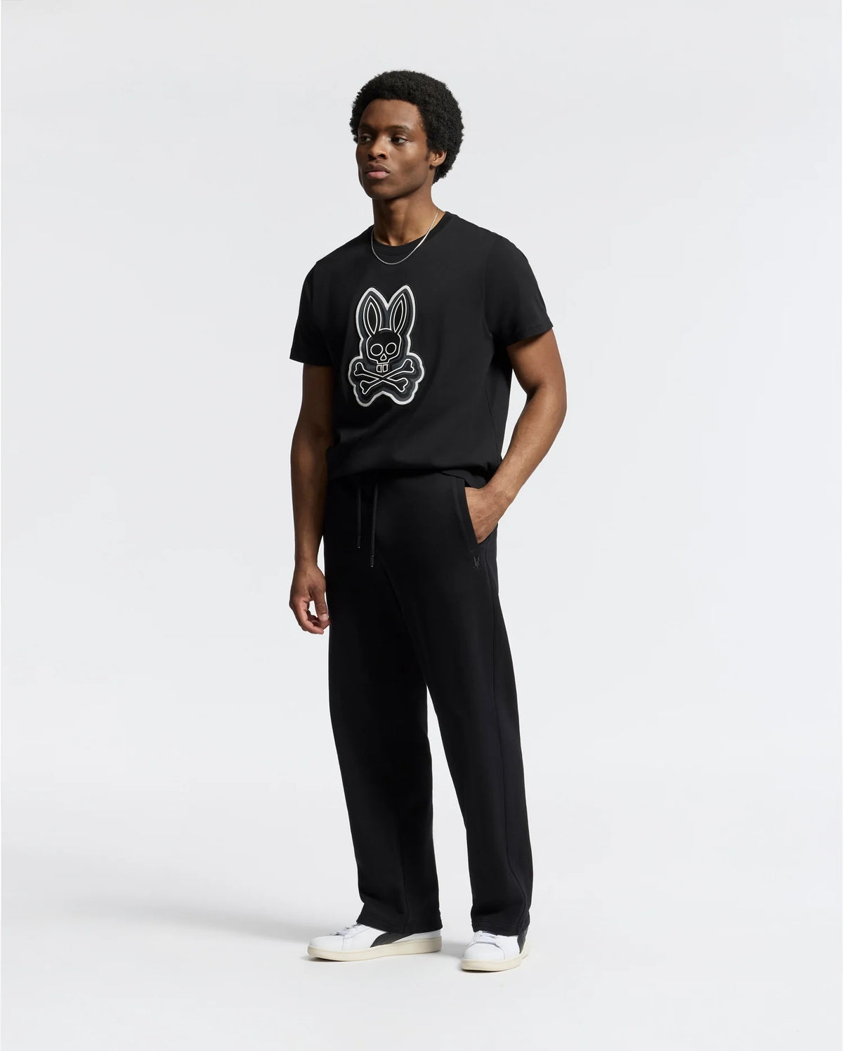 A person in a black T-shirt featuring a stylized rabbit graphic, Psycho Bunny MENS LYNX WIDE LEG CLASSIC FRENCH TERRY SWEATPANT - B6P379E200, and white sneakers stands against a plain background.