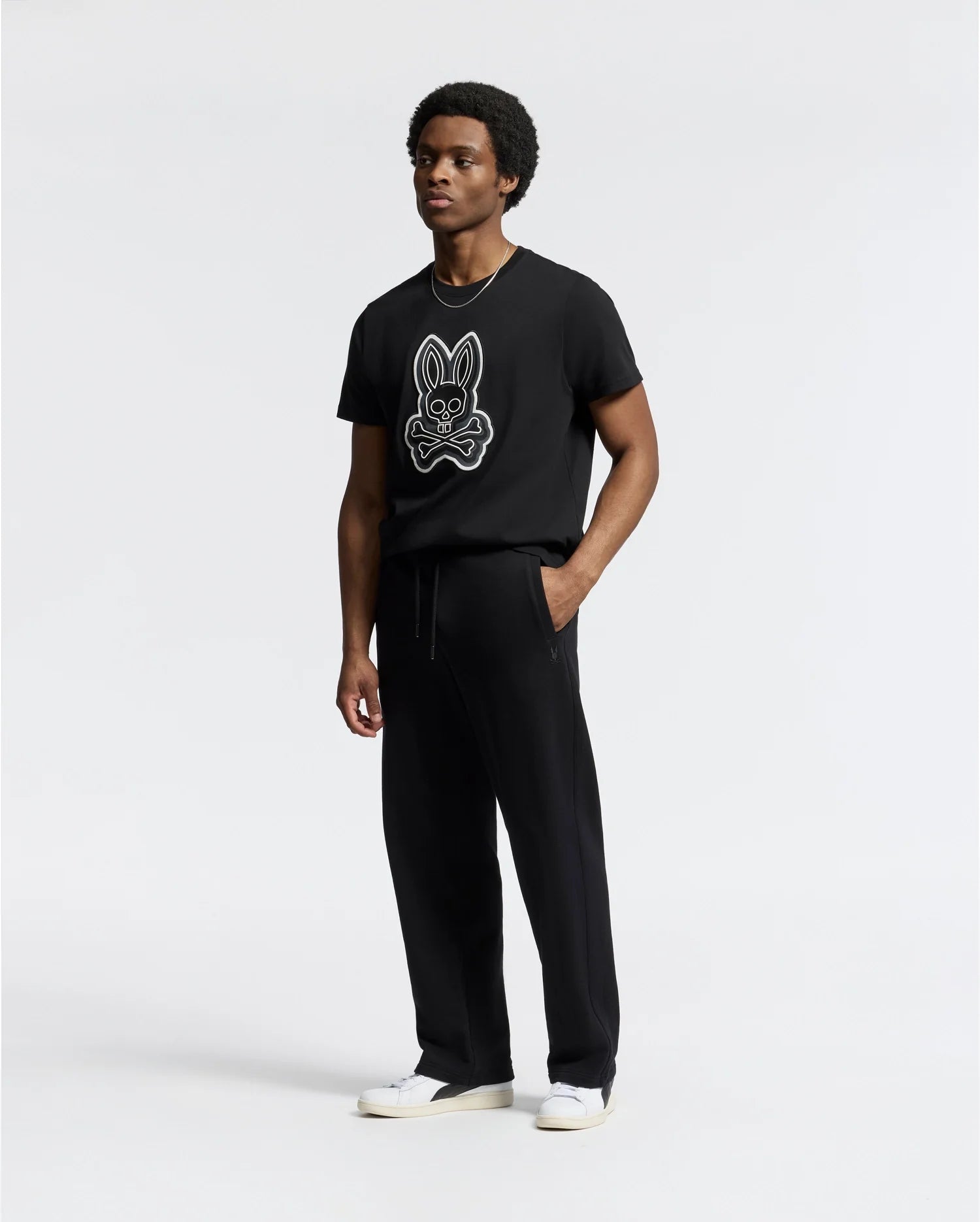 A person in a black T-shirt featuring a stylized rabbit graphic, Psycho Bunny MENS LYNX WIDE LEG CLASSIC FRENCH TERRY SWEATPANT - B6P379E200, and white sneakers stands against a plain background.