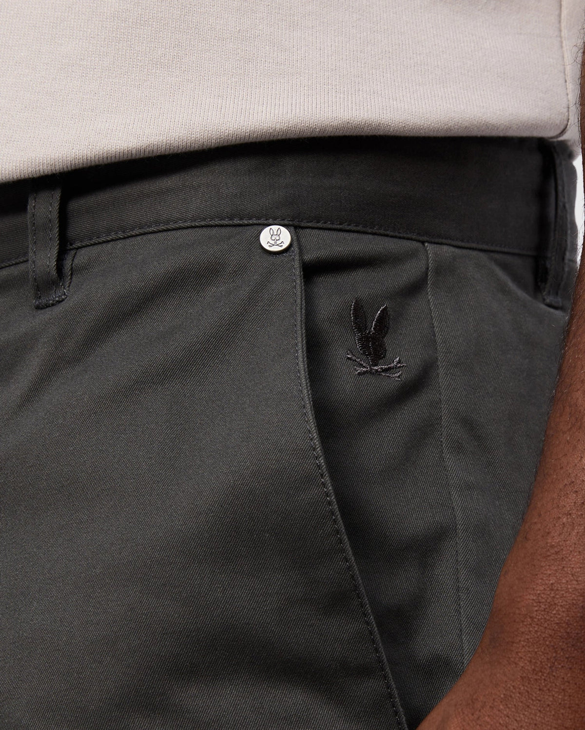 A close-up of a person wearing the Psycho Bunny MENS GILMAN CHINO PANT - B6P393Z1WB in dark green showcases a small black embroidered logo above the pocket. A metallic Psycho Bunny-branded button is visible near the waistband. The stretch fabric offers comfort, and a hint of a beige shirt peeks out above the pants.