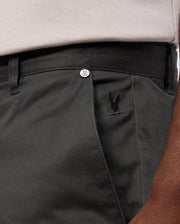 A close-up of a person wearing the Psycho Bunny MENS GILMAN CHINO PANT - B6P393Z1WB in dark green showcases a small black embroidered logo above the pocket. A metallic Psycho Bunny-branded button is visible near the waistband. The stretch fabric offers comfort, and a hint of a beige shirt peeks out above the pants.