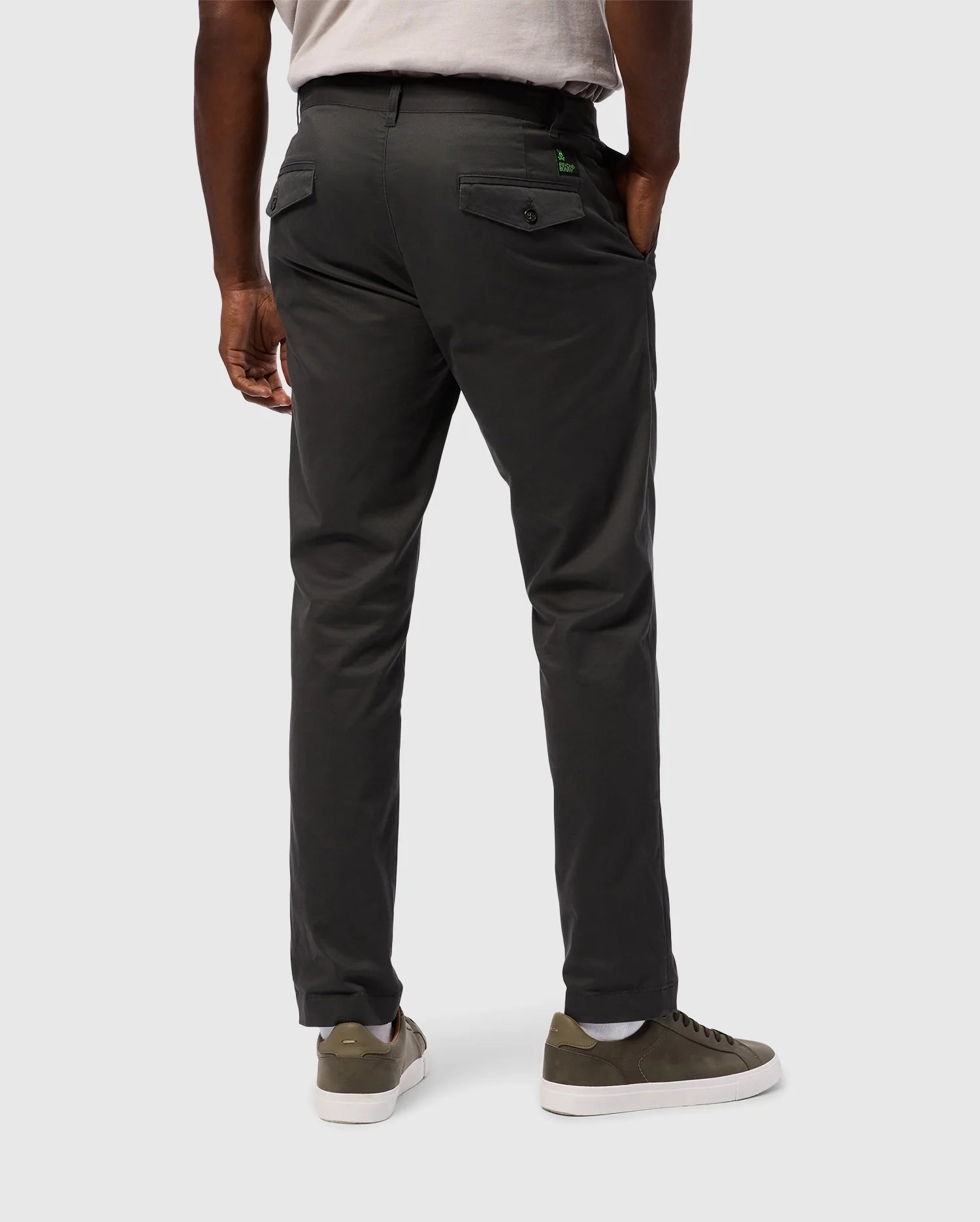 A person is facing away from the camera, wearing Psycho Bunny's MENS GILMAN CHINO PANT - B6P393Z1WB in dark gray, which provides stretch and comfort. They have paired it with a light gray shirt, one hand tucked into their pocket, and are sporting green and white sneakers. The scene is set against a plain white background.