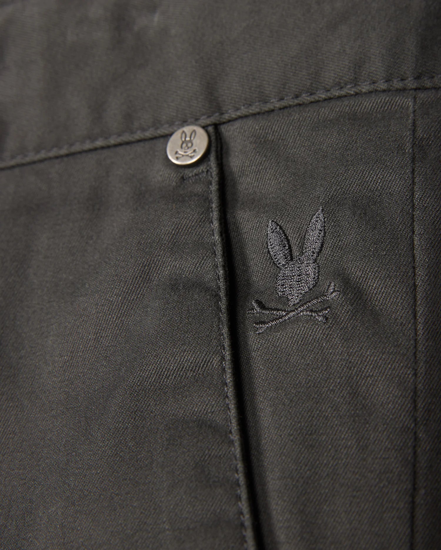 Close-up of the MENS GILMAN CHINO PANT by Psycho Bunny, showcasing a dark chino fabric with a pocket. A small, embroidered rabbit skull and crossbones logo adds unique flair, complemented by a metal button embossed with the same rabbit design. The comfortable stretch material ensures both style and ease of movement.