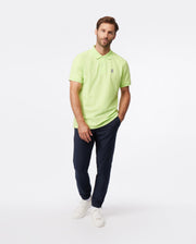 A man stands against a white background, wearing a lime green polo shirt, Psycho Bunny's MENS MADISON COMMUTER PANT - B6P395Z1WB with an elasticized waistband, and white sneakers, looking at the camera with a neutral expression.