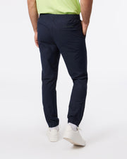 Rear view of a model wearing Psycho Bunny's MEN'S MADISON COMMUTER PANT - B6P395Z1WB joggers with an elasticized waistband, paired with a green t-shirt, standing against a white background. The model's footwear includes casual white sneakers.
