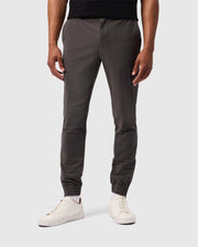 A person in the MENS MADISON COMMUTER PANT - B6P395Z1WB by Psycho Bunny, featuring dark gray jogger pants with elastic cuffs at the ankles and a detachable keychain, stands against a plain gray background in white sneakers. These joggers combine style with practicality. The upper body is not visible.