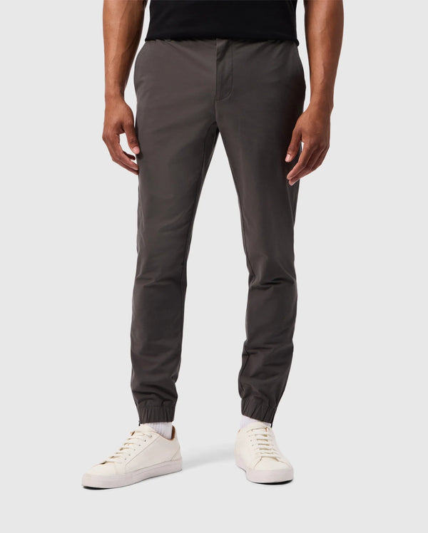 A person in the MENS MADISON COMMUTER PANT - B6P395Z1WB by Psycho Bunny, featuring dark gray jogger pants with elastic cuffs at the ankles and a detachable keychain, stands against a plain gray background in white sneakers. These joggers combine style with practicality. The upper body is not visible.