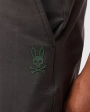 A close-up showcases the Psycho Bunny MENS MADISON COMMUTER PANT - B6P395Z1WB, made from dark fabric with a distinctive green rabbit skull and crossbones emblem. A detachable Bunny keychain dangles nearby, adding character to the design, while zippered expansion vents suggest versatility. A person's arm partially appears on the right.