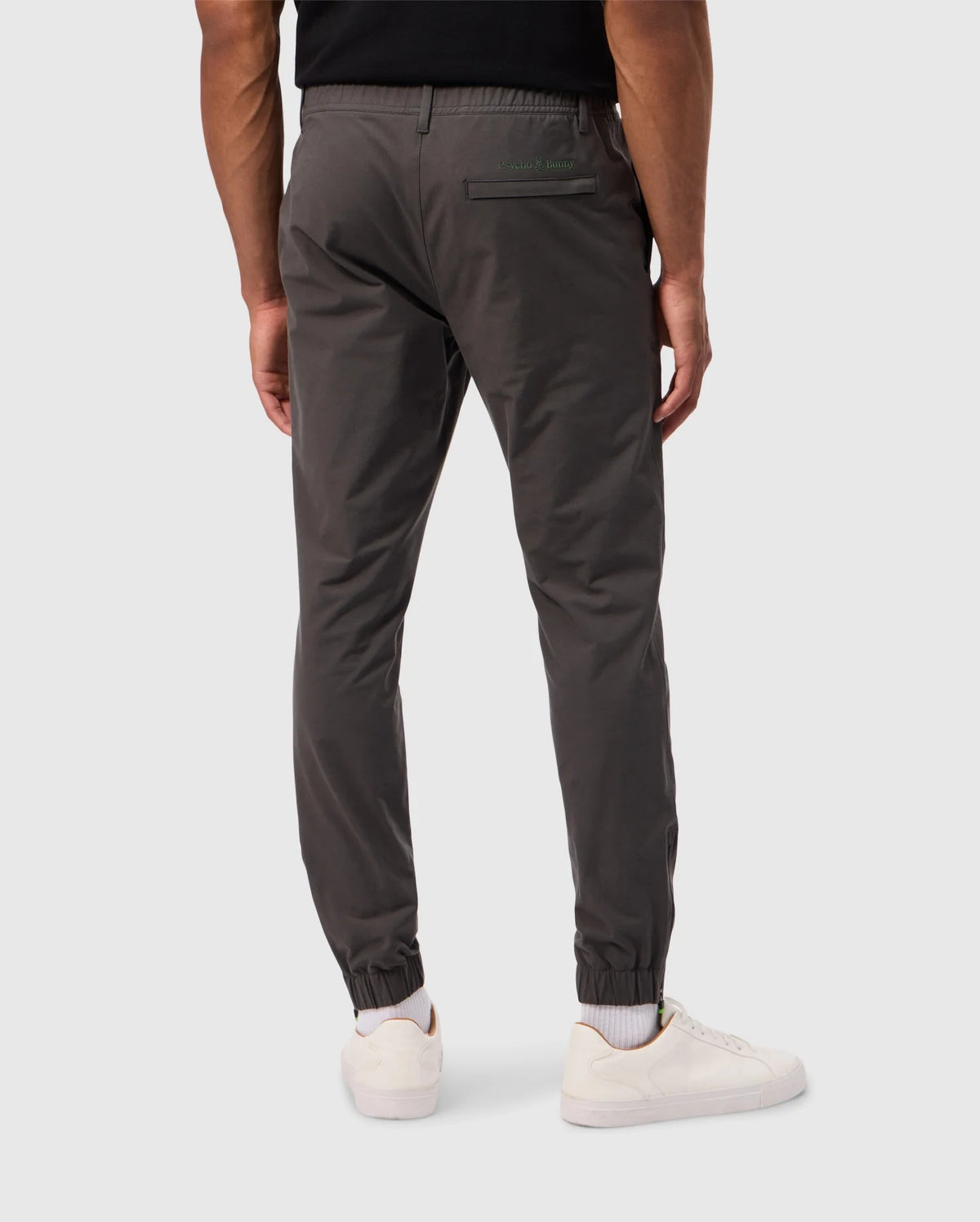 A person wearing the MENS MADISON COMMUTER PANT - B6P395Z1WB by Psycho Bunny, styled in dark gray jogger fashion, and white sneakers stands against a light gray background. These commuter pants are designed with an elastic waistband, cuffs, a single back pocket showcasing a green label, and include a detachable keychain for added convenience.