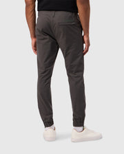A person wearing the MENS MADISON COMMUTER PANT - B6P395Z1WB by Psycho Bunny, styled in dark gray jogger fashion, and white sneakers stands against a light gray background. These commuter pants are designed with an elastic waistband, cuffs, a single back pocket showcasing a green label, and include a detachable keychain for added convenience.