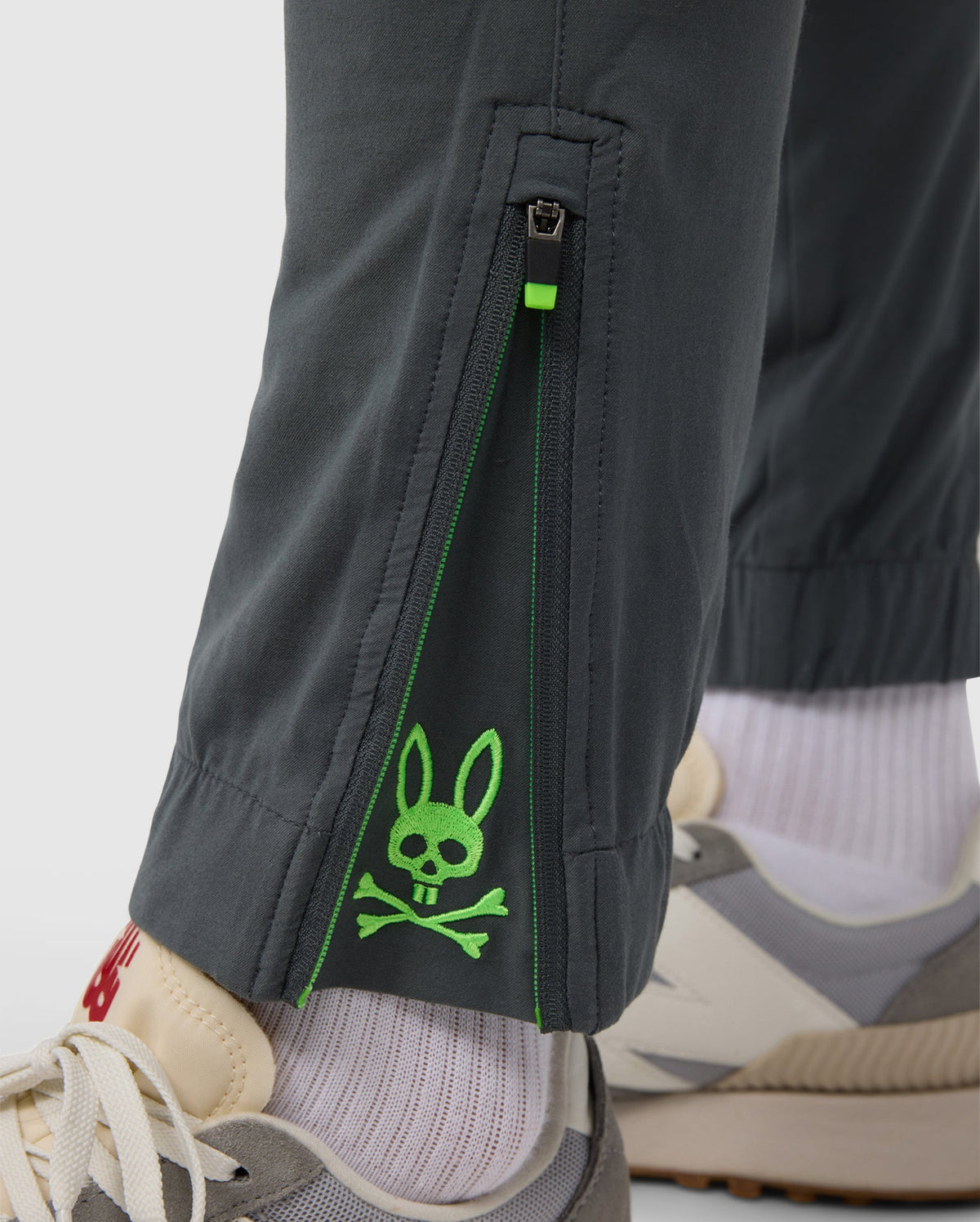 Close-up of a person's lower leg showing a grey Psycho Bunny MEN'S MADISON COMMUTER PANT - B6P395Z1WB leg with a green zipper and neon green embroidered Bunny logos. The person is wearing white socks and beige sneakers.