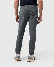 A person facing away, wearing Psycho Bunny's MENS MADISON COMMUTER PANT - B6P395Z1WB with an elasticized waistband and white sneakers, shown from the waist down against a plain white background. The pants have a small green tag near the