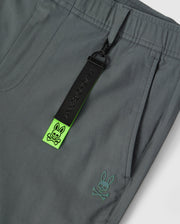 Close-up image of gray men's Madison commuter pant by Psycho Bunny featuring a black zip pull with the word "badbunny" and a neon green strap with embroidered Bunny logos.