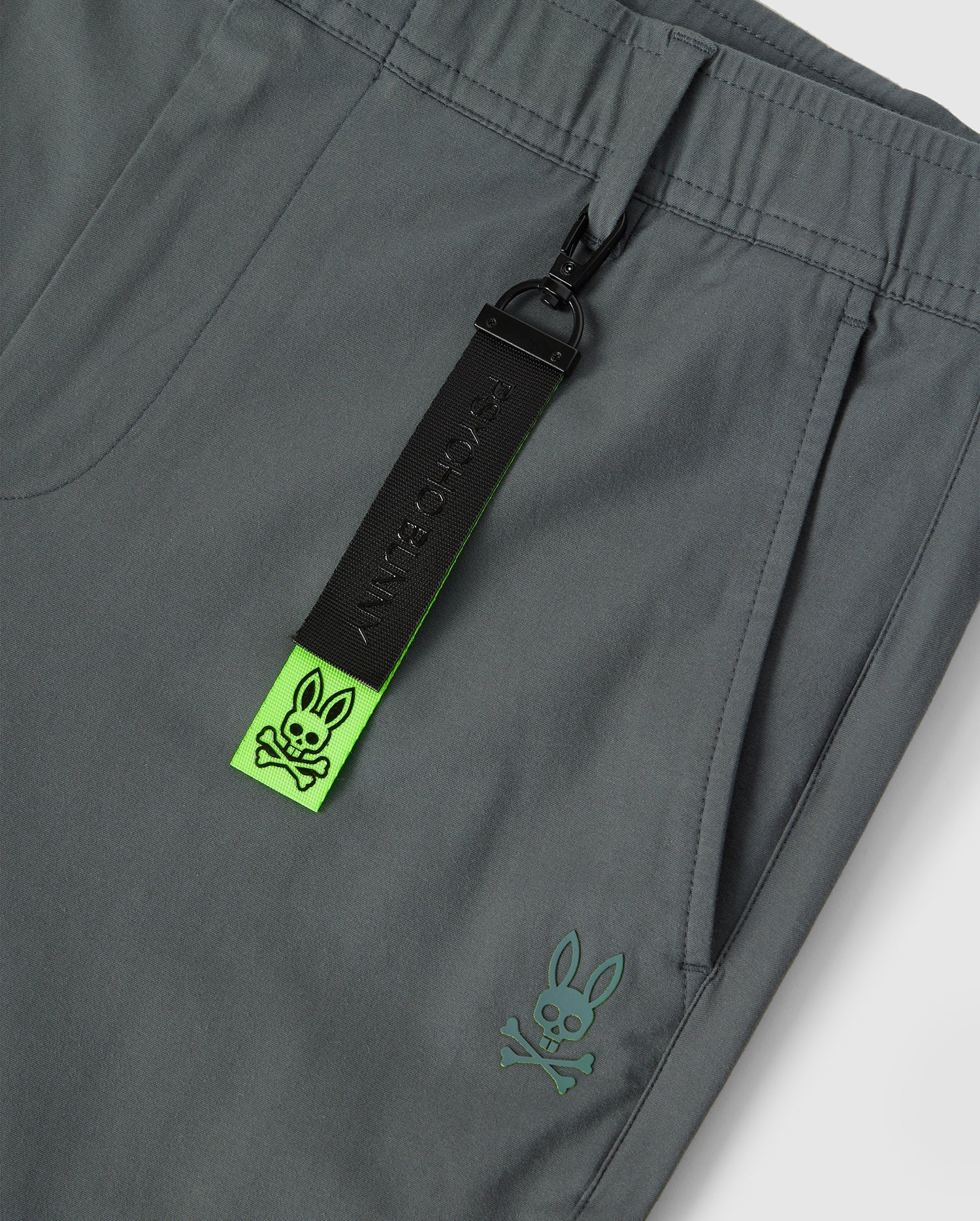 Close-up image of gray men's Madison commuter pant by Psycho Bunny featuring a black zip pull with the word "badbunny" and a neon green strap with embroidered Bunny logos.