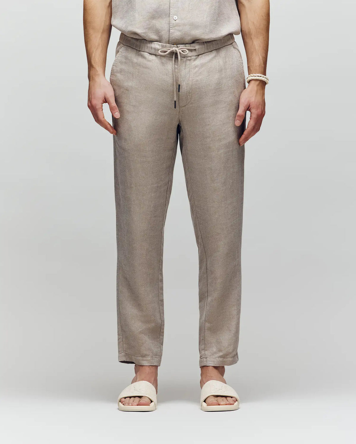 A person wears Psycho Bunny MENS FATE LINEN DRAWSTRING PANT (B6P471C200) and cream slide sandals, standing with hands relaxed at their sides against a plain light background.