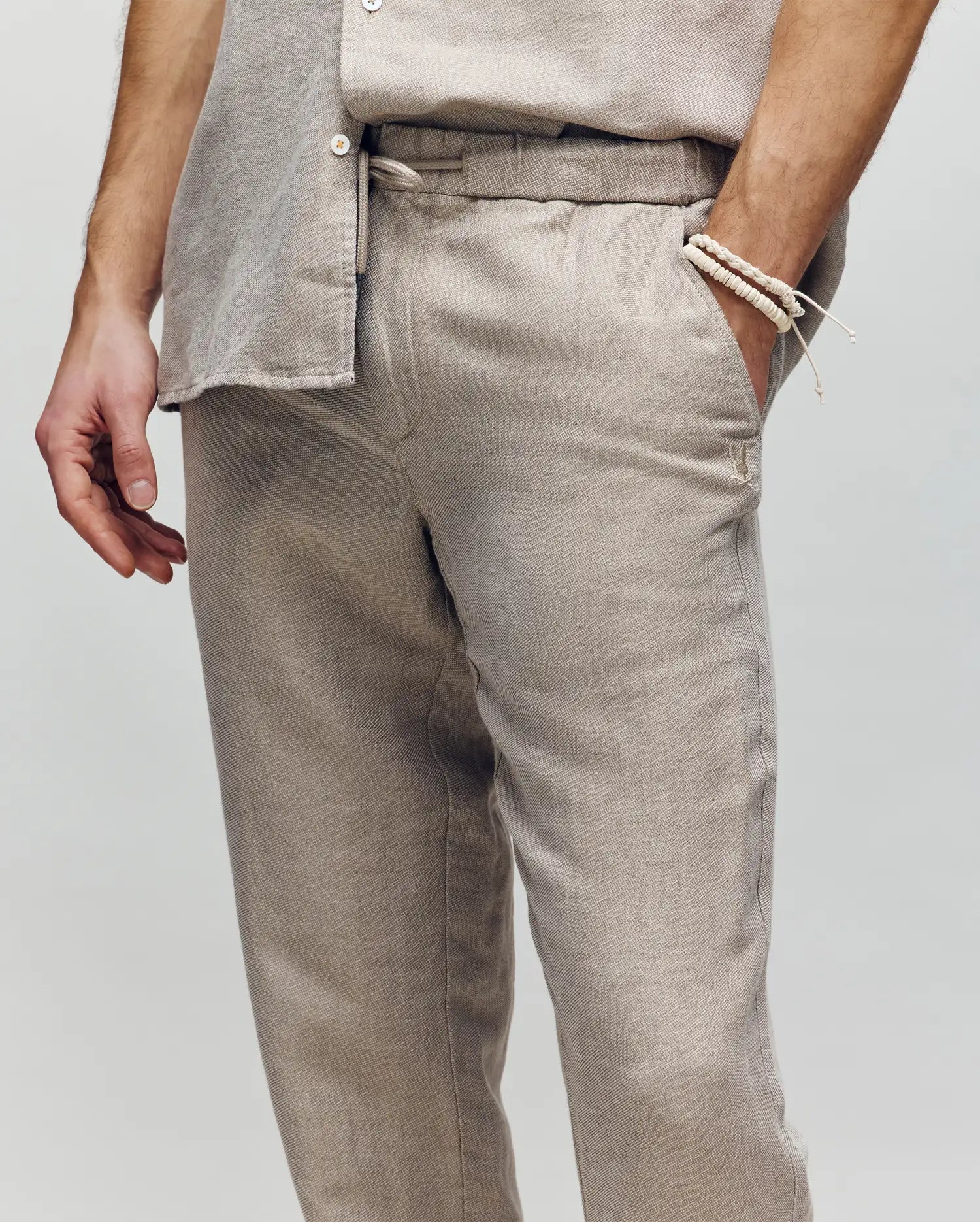 A person wears Psycho Bunny’s MENS FATE LINEN DRAWSTRING PANT (B6P471C200) in light beige linen blend, paired with a matching button-up shirt and a beige beaded bracelet. They stand with one hand in their pocket against a neutral background.
