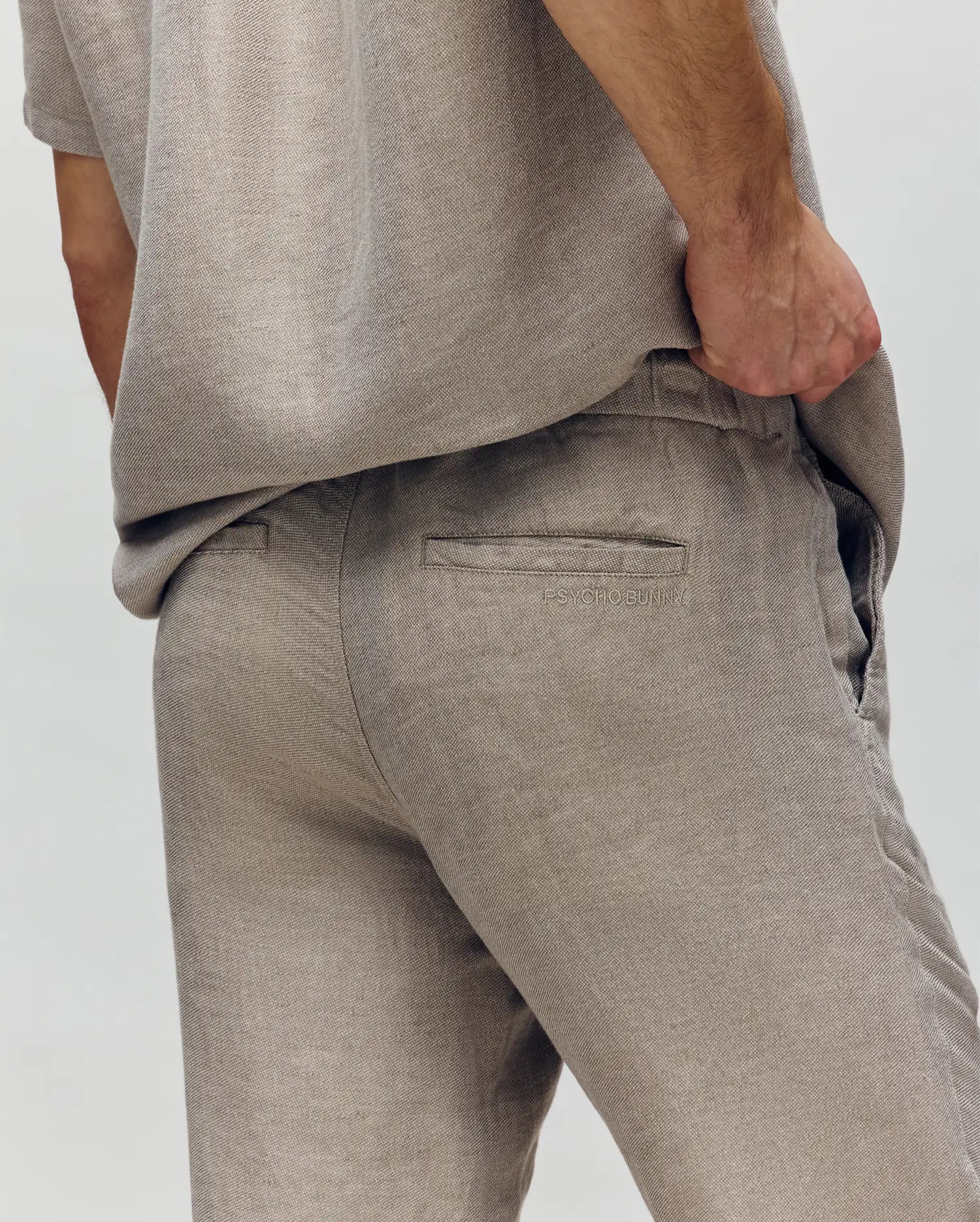 A person models the Psycho Bunny MENS FATE LINEN DRAWSTRING PANT (B6P471C200) in beige, highlighting the back pocket and textured linen blend for cooling comfort. Their left hand rests on their hip against a plain, neutral background.