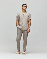 A man wearing sunglasses, a short-sleeve beige button-up shirt, and Psycho Bunny’s MENS FATE LINEN DRAWSTRING PANT (B6P471C200) with matching sandals stands against a light gray background, hands in pockets, looking confidently to the side.