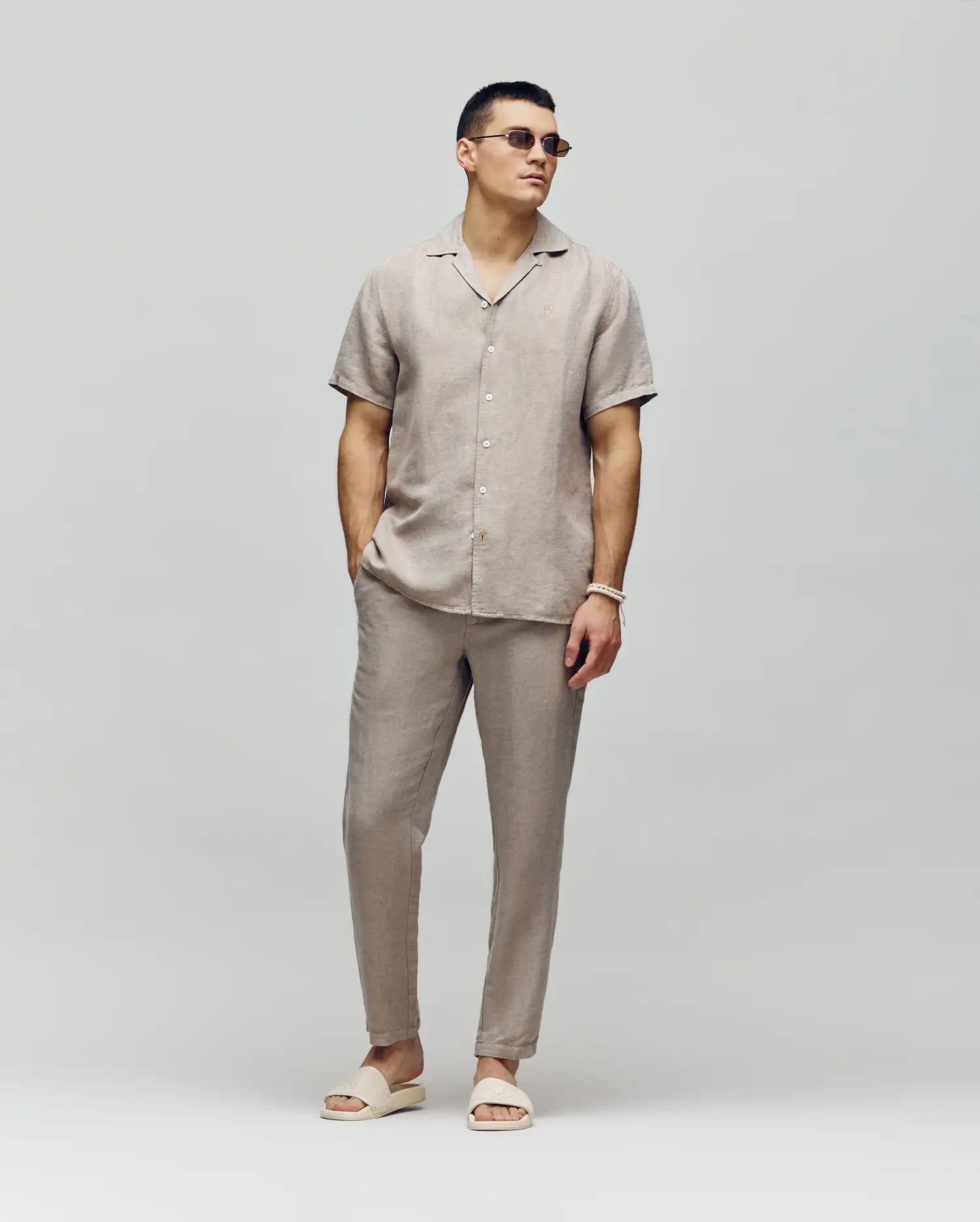 A man wearing sunglasses, a short-sleeve beige button-up shirt, and Psycho Bunny’s MENS FATE LINEN DRAWSTRING PANT (B6P471C200) with matching sandals stands against a light gray background, hands in pockets, looking confidently to the side.