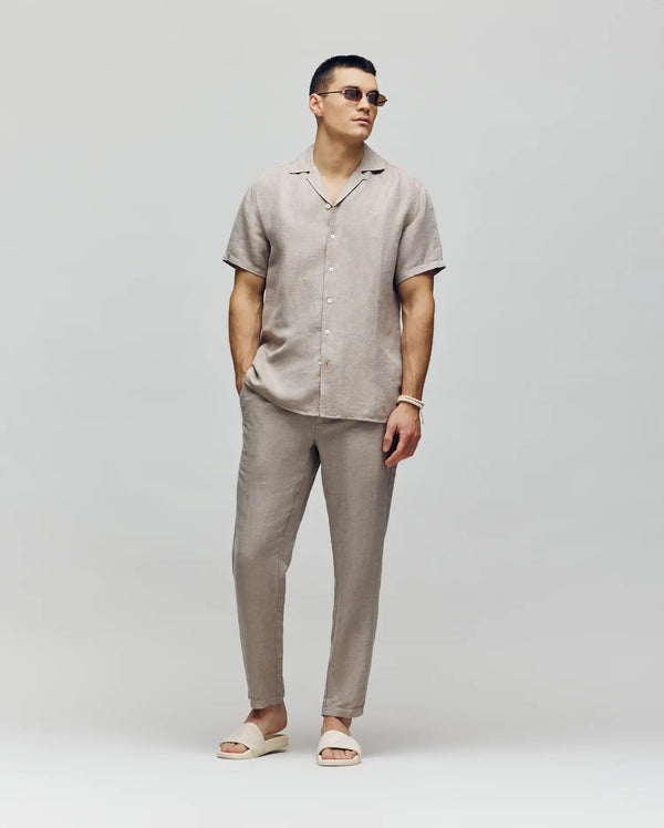 A man wearing sunglasses, a short-sleeve beige button-up shirt, and Psycho Bunny’s MENS FATE LINEN DRAWSTRING PANT (B6P471C200) with matching sandals stands against a light gray background, hands in pockets, looking confidently to the side.