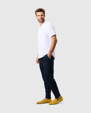 A man with short light brown hair stands relaxed, looking to the right. He is wearing a white short-sleeve shirt, Psycho Bunny dark blue MENS FATE LINEN DRAWSTRING PANT - B6P471C200 trousers, and bright yellow loafers. The background is plain light gray.