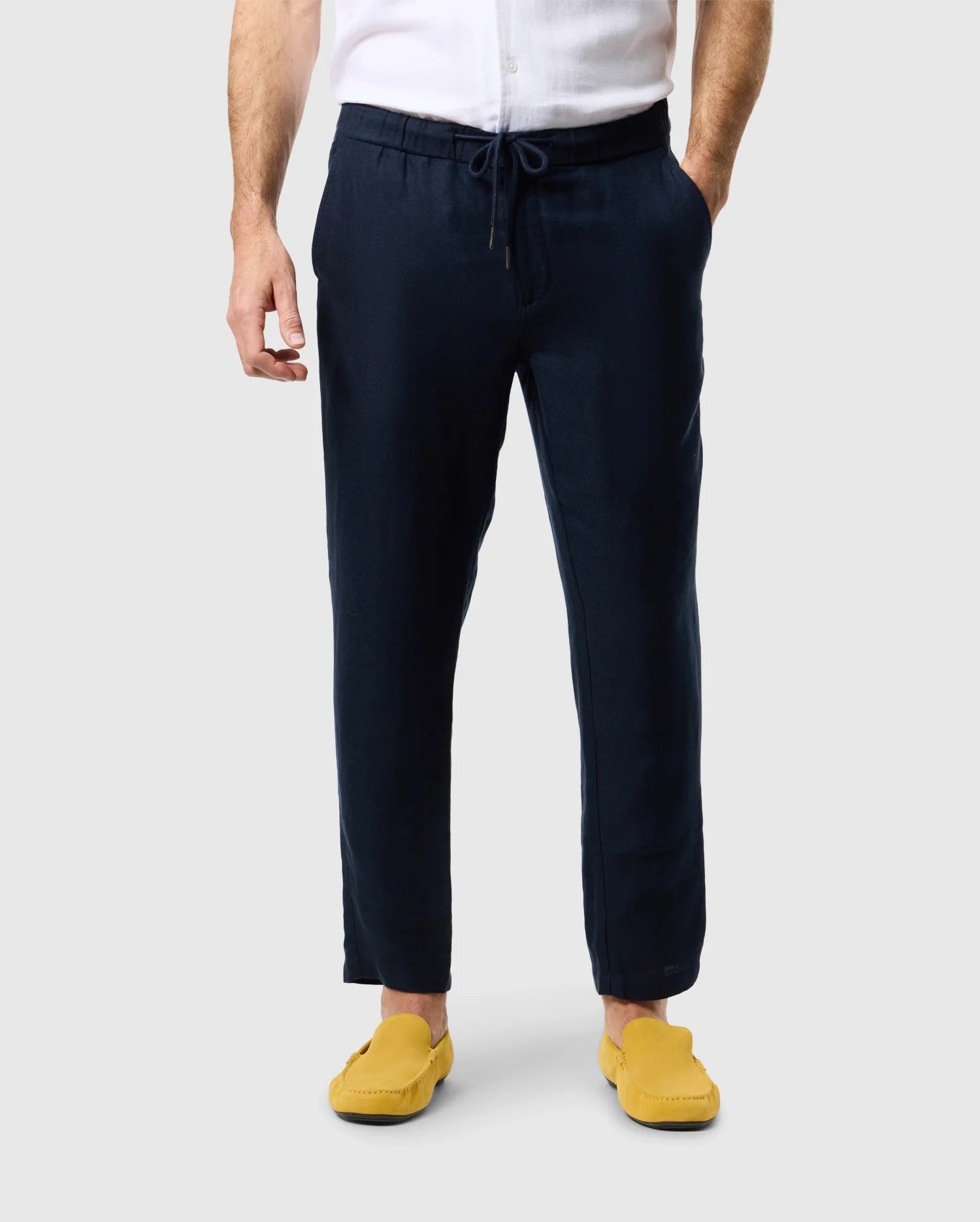 Against a plain light background, a person stands wearing a white button-down shirt paired with Psycho Bunny's MENS FATE LINEN DRAWSTRING PANT - B6P471C200 in navy blue and yellow loafers. The focus is on the lower half of their body, highlighting the summer trousers and footwear.