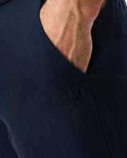Close-up of a person's hand placed in the pocket of Psycho Bunny's MENS FATE LINEN DRAWSTRING PANT - B6P471C200 in navy blue. The pants, featuring a small embroidered emblem near the pocket, appear to be soft and comfortable—perfect for summer trousers.