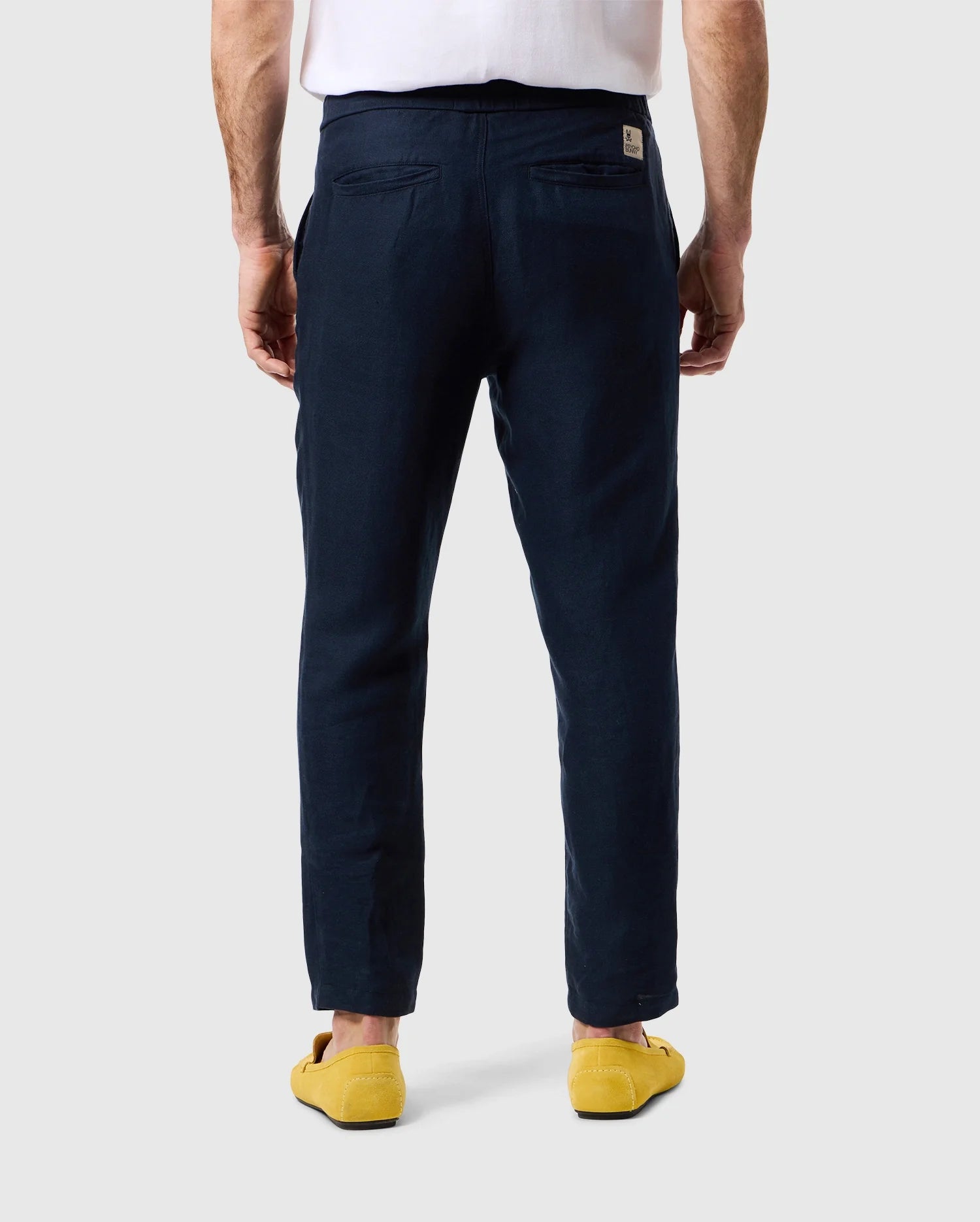 A man in a white shirt and Psycho Bunny MENS FATE LINEN DRAWSTRING PANT - B6P471C200, stands with his back facing the camera, hands down by his sides. He is wearing bright yellow shoes. The background is plain and light gray.