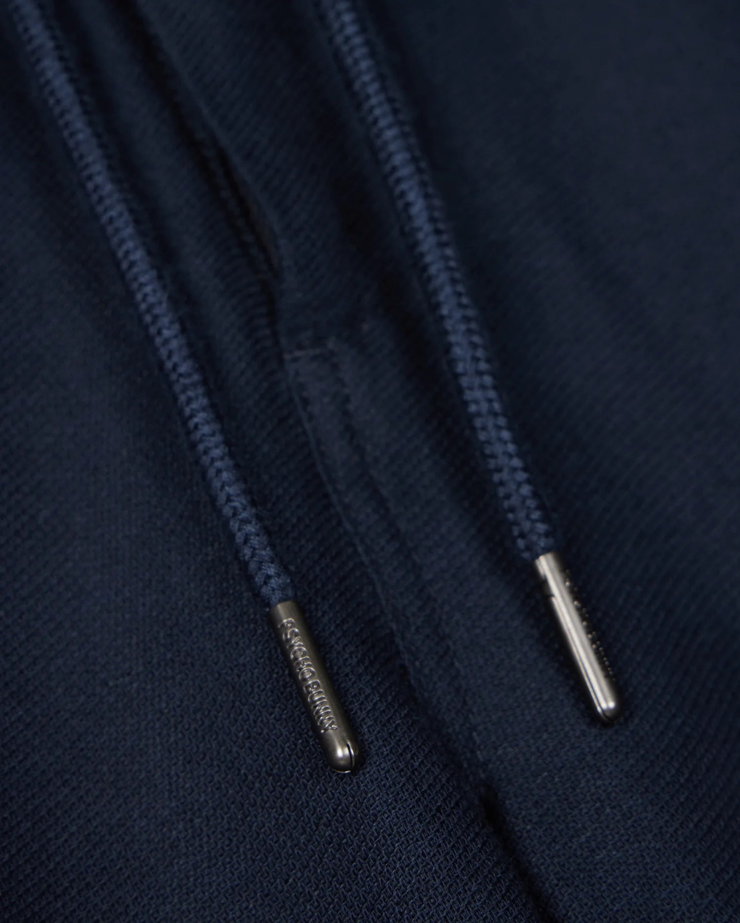 Close-up of navy blue drawstrings with silver metal tips on a dark fabric background, perfect for the Psycho Bunny MENS FATE LINEN DRAWSTRING PANT - B6P471C200. The metal tips have small engraved text, but the content is not fully readable in the image. Ideal for those summer trousers that need both style and durability.