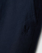 A close-up of the dark blue fabric from the Psycho Bunny MENS FATE LINEN DRAWSTRING PANT - B6P471C200 showcases a small, embroidered bunny logo with crossbones underneath, located near the pocket seam. The visible texture of the fabric gives it a cozy appearance, making it perfect for your next pair of men’s linen pants.