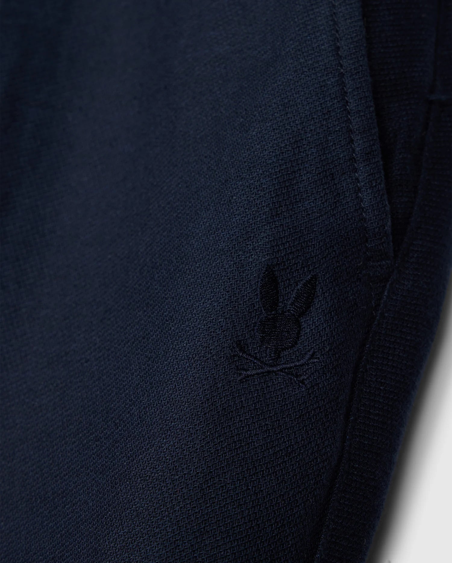 A close-up of the dark blue fabric from the Psycho Bunny MENS FATE LINEN DRAWSTRING PANT - B6P471C200 showcases a small, embroidered bunny logo with crossbones underneath, located near the pocket seam. The visible texture of the fabric gives it a cozy appearance, making it perfect for your next pair of men’s linen pants.