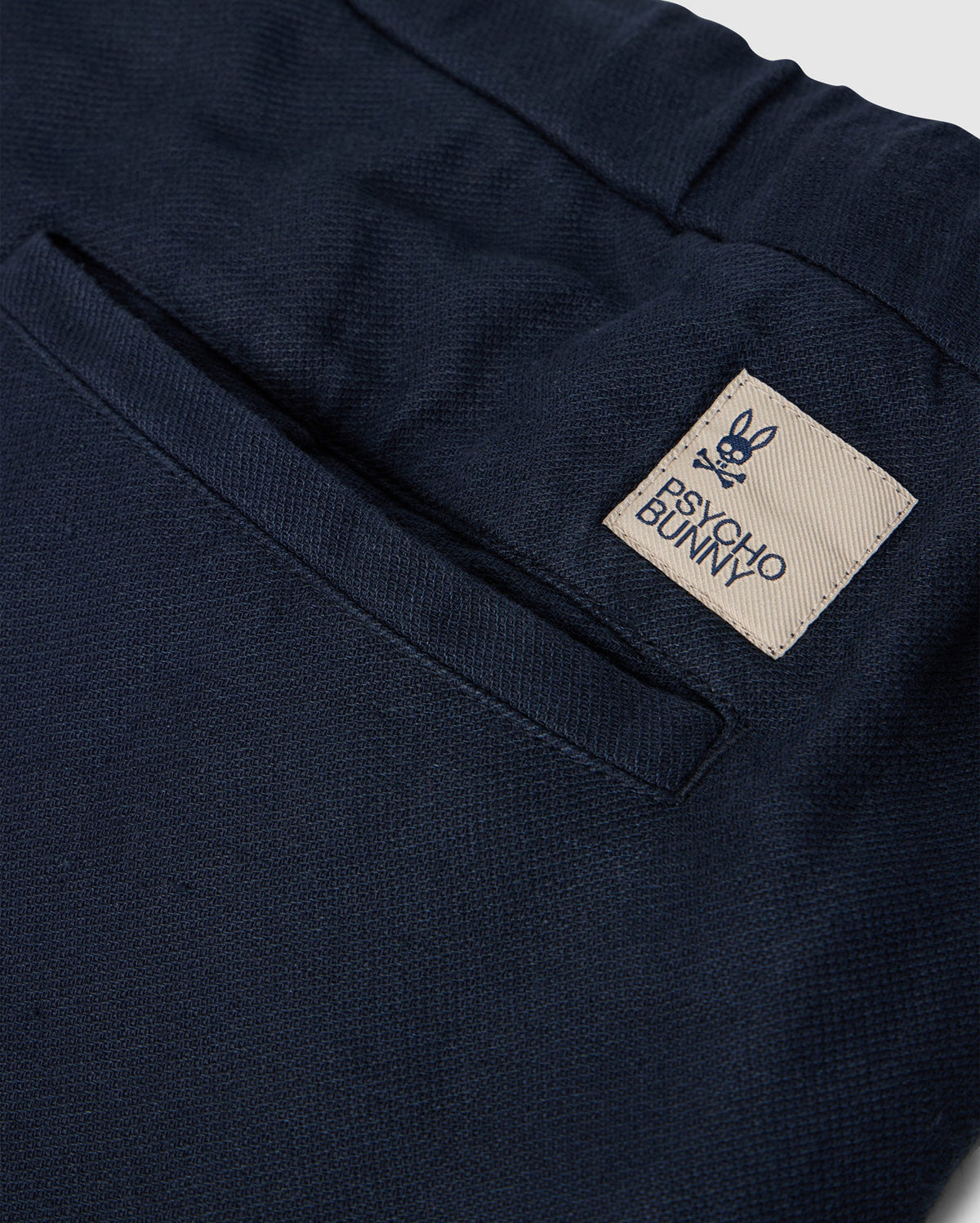 Close-up of the back pocket of MENS FATE LINEN DRAWSTRING PANT - B6P471C200 by Psycho Bunny. The black summer trousers feature a beige tag with a bunny face logo and crossbones underneath, along with the text "Psycho Bunny.