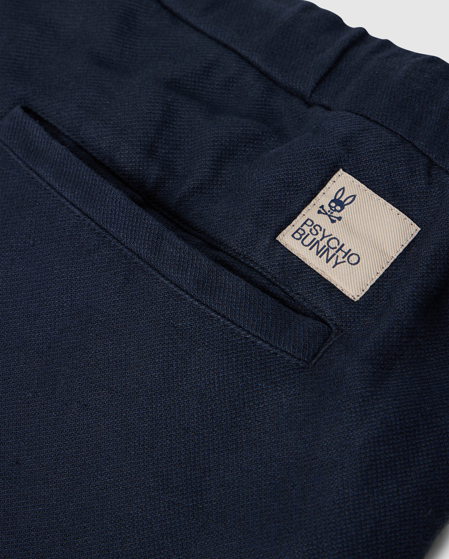 Close-up of the back pocket of MENS FATE LINEN DRAWSTRING PANT - B6P471C200 by Psycho Bunny. The black summer trousers feature a beige tag with a bunny face logo and crossbones underneath, along with the text "Psycho Bunny.