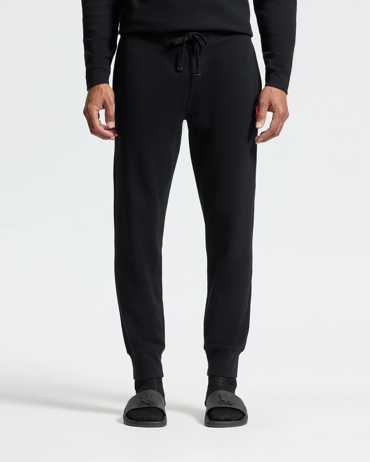 A person is shown wearing the Psycho Bunny MENS VAIL WAFFLE LOUNGE PANT in black, paired with black slide sandals, standing against a plain white background. The focus is on the lower half of their body, showcasing the pants' drawstring and relaxed fit ideal for lounging.