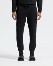 A person is shown wearing the Psycho Bunny MENS VAIL WAFFLE LOUNGE PANT in black, paired with black slide sandals, standing against a plain white background. The focus is on the lower half of their body, showcasing the pants' drawstring and relaxed fit ideal for lounging.