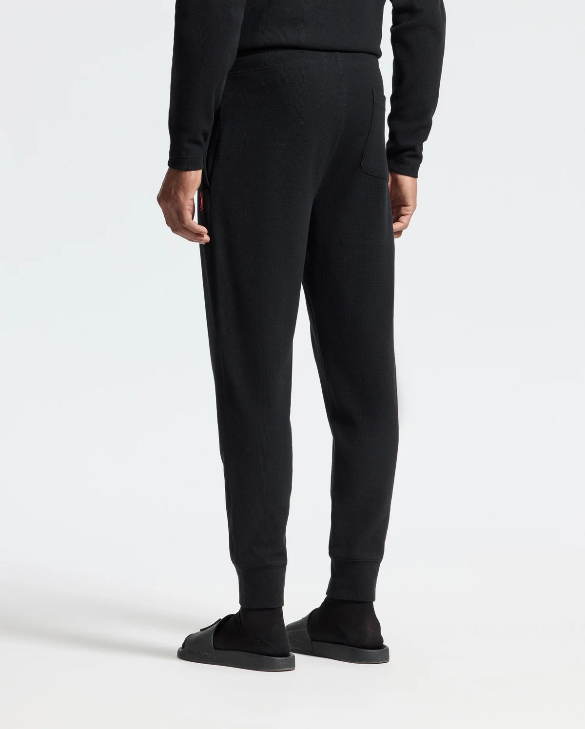 A person wearing the Psycho Bunny MENS VAIL WAFFLE LOUNGE PANT - B6P673E200 in black, possibly made from a cozy Pima cotton blend, and black shoes stands with their back to the camera against a plain white background.