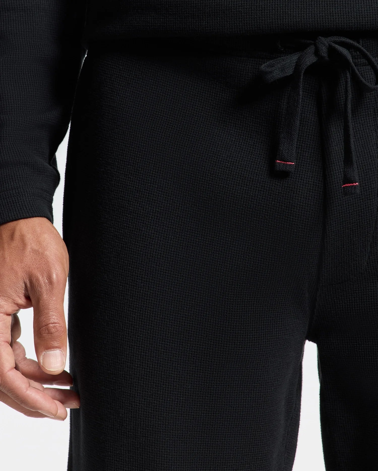 A person is wearing a black textured long-sleeve top paired with Psycho Bunny's Men's Vail Waffle Lounge Pants (B6P673E200), which feature a drawstring with red accents. The focus is on the lower part of the outfit and the person's hand, set against a plain white backdrop.