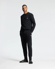 A man stands against a plain background wearing a black sweatshirt paired with Psycho Bunny's MENS VAIL WAFFLE LOUNGE PANT - B6P673E200, featuring a small red logo. His hands are tucked into the pockets of these joggers, crafted from a soft pima cotton blend, and he finishes off the relaxed look with black slides.