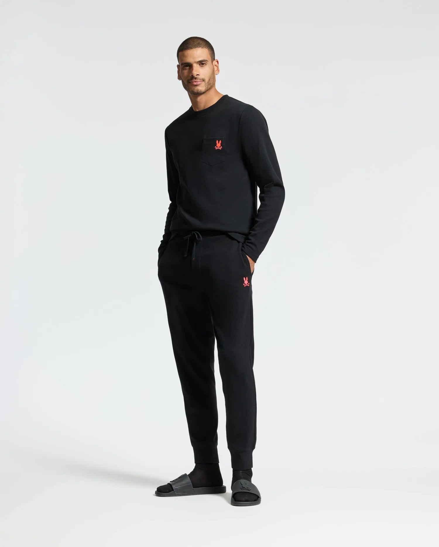 A man stands against a plain background wearing a black sweatshirt paired with Psycho Bunny's MENS VAIL WAFFLE LOUNGE PANT - B6P673E200, featuring a small red logo. His hands are tucked into the pockets of these joggers, crafted from a soft pima cotton blend, and he finishes off the relaxed look with black slides.