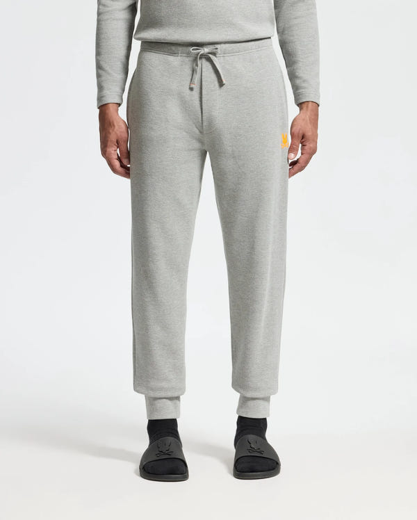 A person wearing the light gray Psycho Bunny MENS VAL WAFFLE LOUNGE PANT - B6P673E200 and a matching top stands with hands at their sides. The pants feature a drawstring waist and have a small yellow logo on the upper right leg. Black socks with a subtle design are visible, all set against a plain white background that enhances the outfit's simplicity.